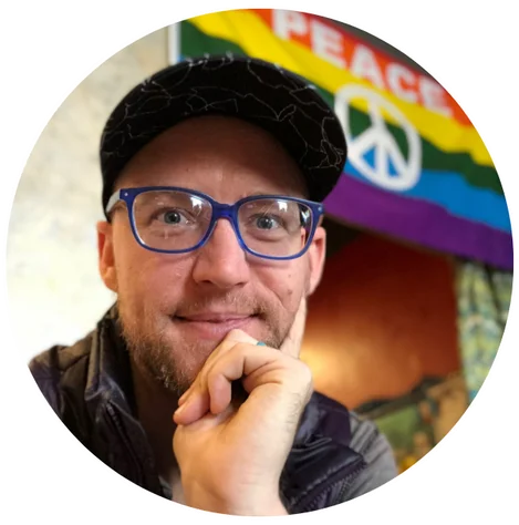 Daniel McCall in his blue glasses, smiling in front of a PEACE flag